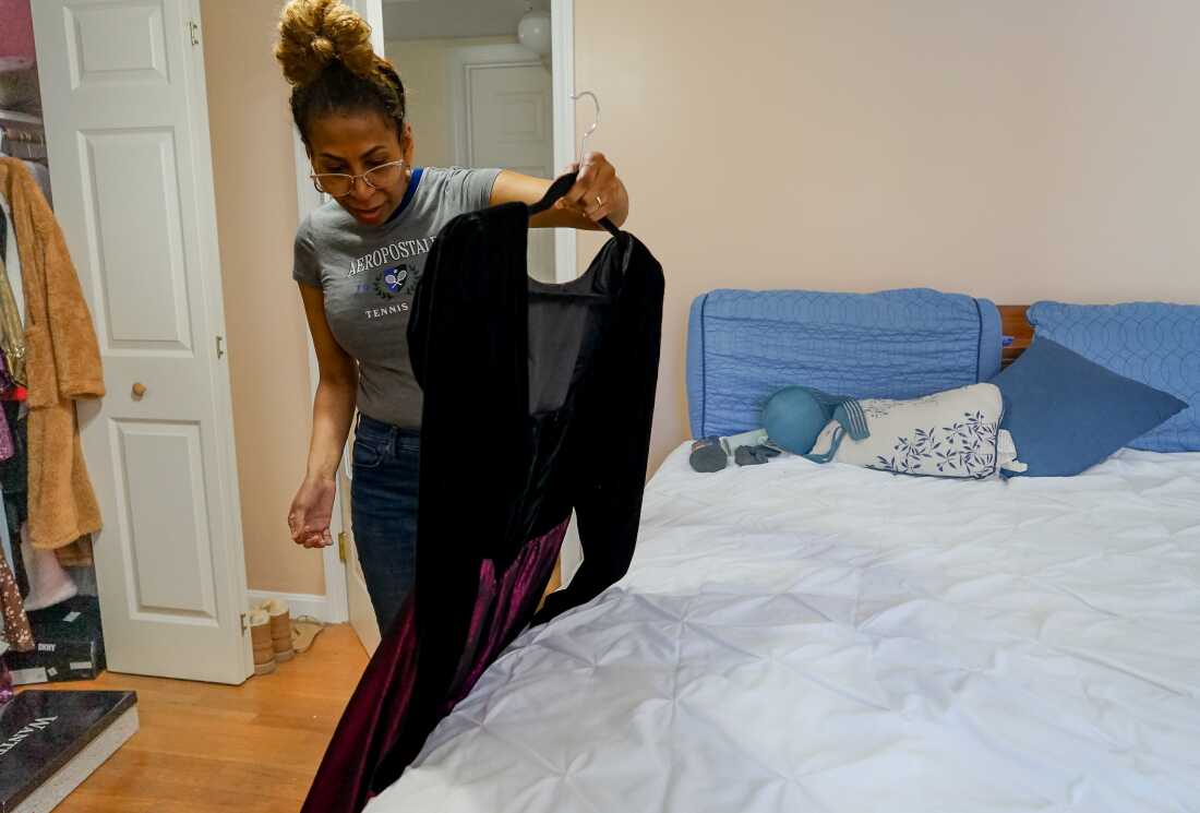 Debra Ragoonanan stands in a bedroom, looking at a long black formal dress on a hanger.