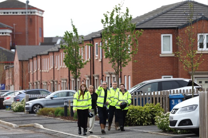 Five people, four of them in high-vis jackets, walk up a street, past a row of houses and parked cars