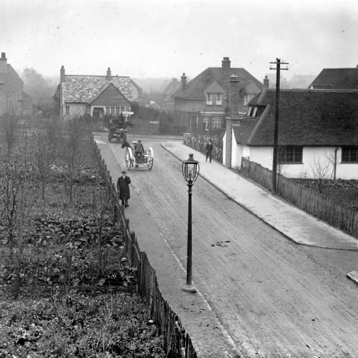 A black and white photo from 1912 shows a horse-drawn cart on an empty street with a thatched cottage on one side and bigger houses in the background