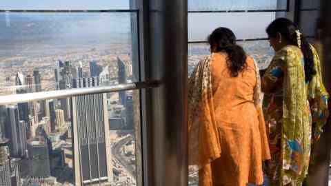 Two Indian tourists, dressed in colorful traditional attire, look out from a high observation deck in the Burj Khalifa, Dubai. The cityscape below features numerous skyscrapers and buildings.