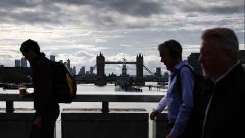 Commuters walk across London Bridge with the Tower Bridge and the HMS Belfast visible in the background.