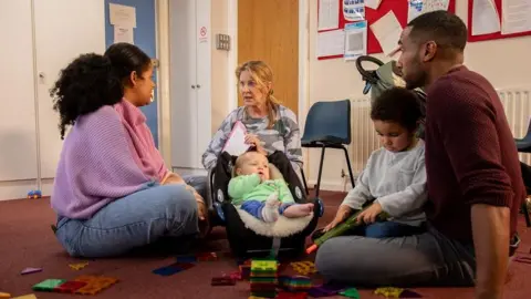 Getty Images A group of adults with their children having a meeting with a childcare worker. They are in a community centre surrounded by toys. 