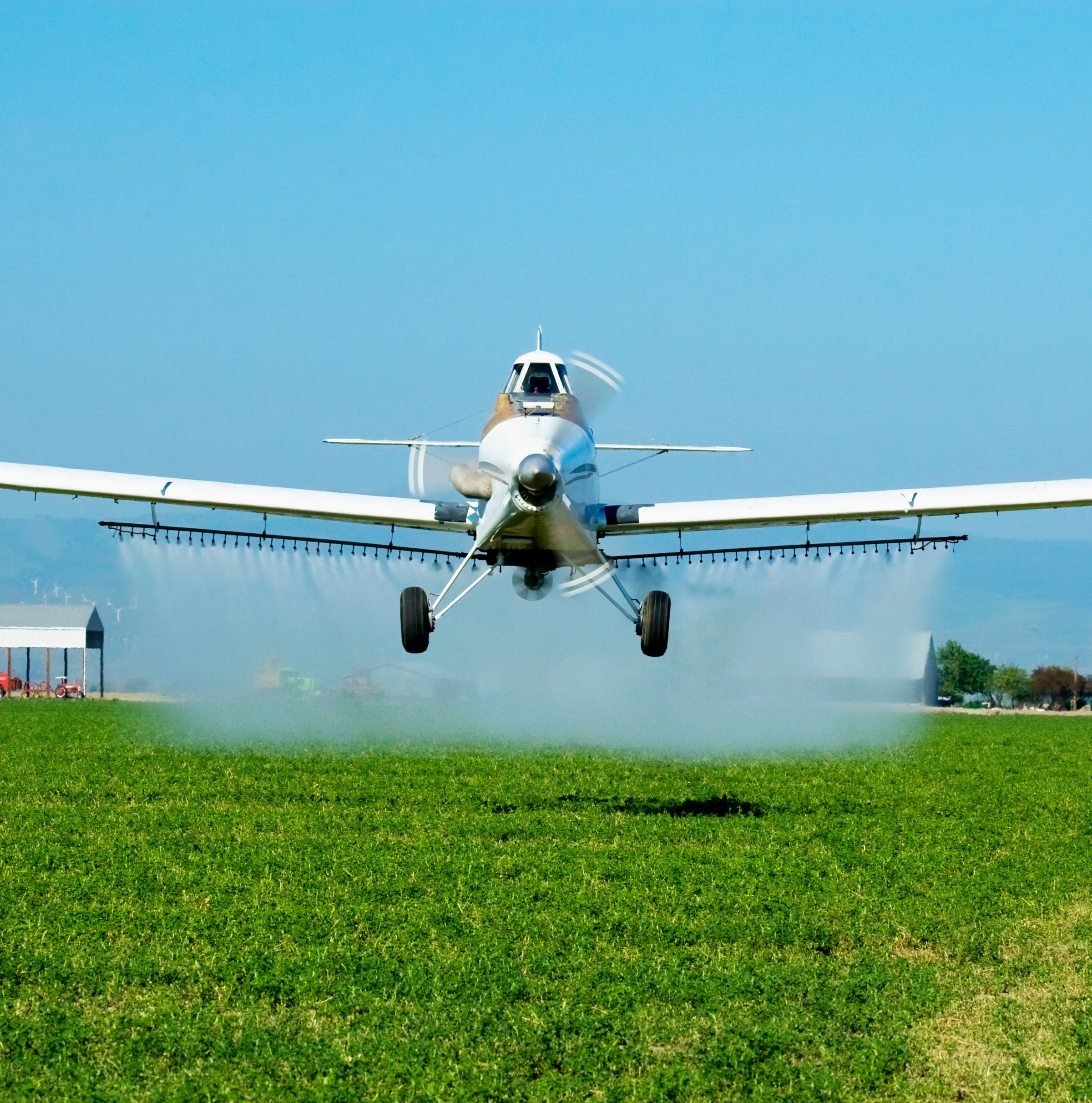 A small twin-engine white plane with crop-dusting equipment under its wings flies slow over a green field, clouds of chemicals descending behind it.