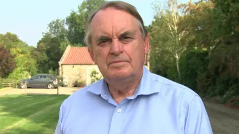 BBC Head-and-shoulders shot of Lord Kirkhope who has receding mousey-brown hair, with grey sections by his temples, and is wearing a light blue shirt. He is standing in a lawned area with trees and shrubs on the right. There is a stone outbuilding in the background with a grey car parked next to it.