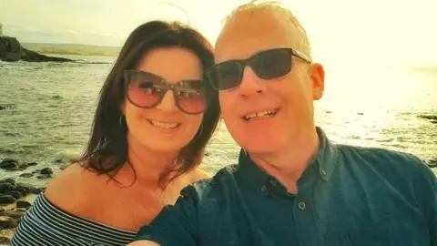 Larry Lowe Larry and his wife pictured by the seaside both are wearing sunglasses and smiling into the camera with the sun shining behind them.