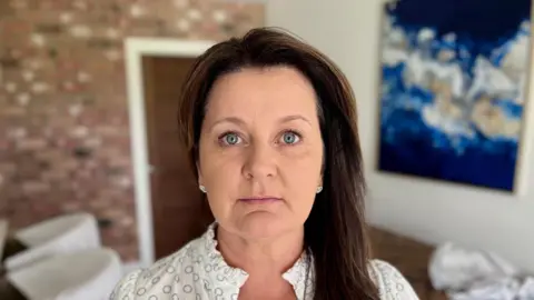 Gini, a woman with shoulder-length brown hair, wearing a white shirt looking directly into the camera