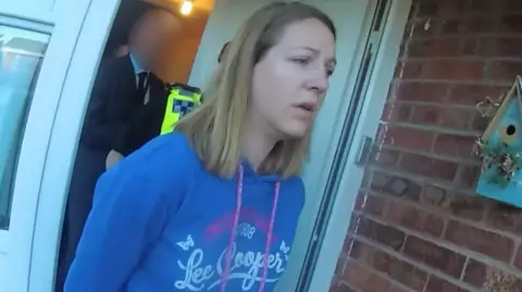 Cheshire Constabulary Police body-cam footage of Lucy Letby being led out of her front door by officers, wearing a blue hoodie with her hands behind her back. 