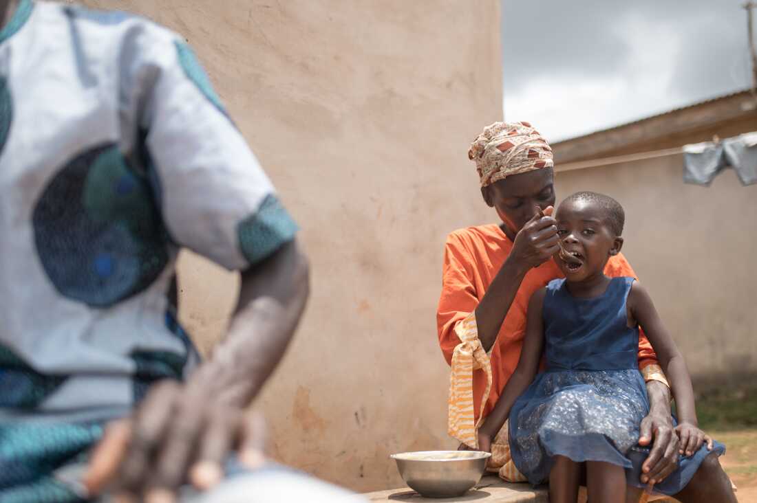 In this photo, Toyin Salami is seated with her young daughter in her lap and is spooning food into the daughter's open mouth. Salami's husband's forearm appears on the left side of the photo.