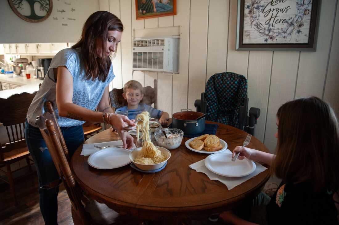 This photo shows two of Caitlyn Kelly's children seated in front of empty plates at a wooden table while she stands at the table, serving them spaghetti from a bowl. A plate with slices of garlic bread sits on the table. 