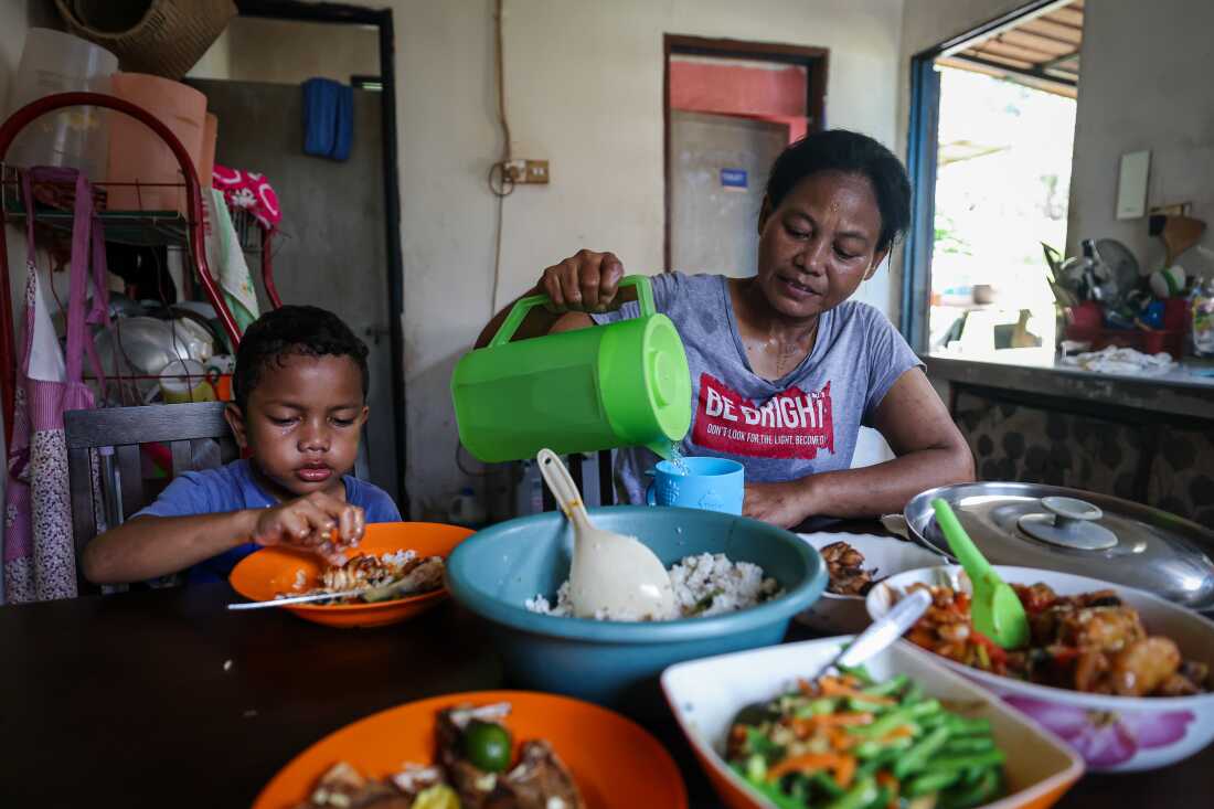 In this photo, Rosnah and her 5-year-old son are seated at a table that has a bowl of rice and several bowls of vegetables on it. Rosnah is pouring water from a pitcher into a drinking cup. Daniel is grasping food from his bowl.