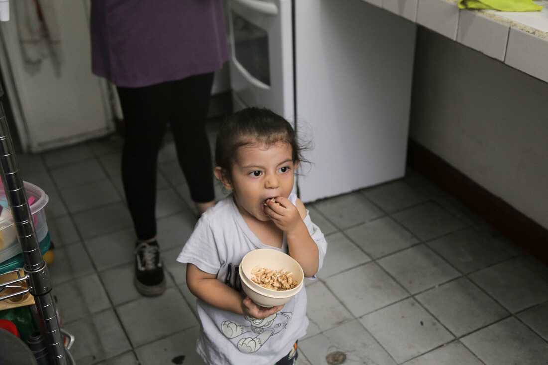 In this photo, 2-year-old Tomás stands in the kitchen while holding a plastic bowl with puffed rice cereal in it. He is using one hand to put some of the cereal into his month.