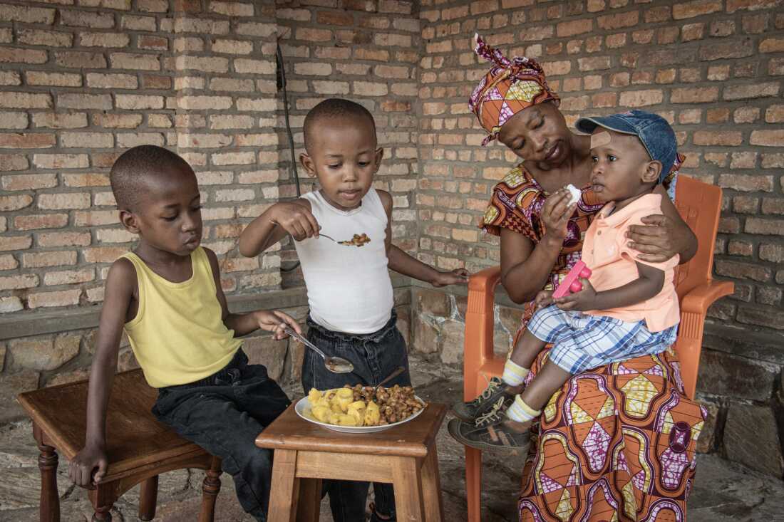 In this photo, Jeannette Uwimbabazi is feeding her children. She is seated with her youngest child, age 2, in her lap. A plate of beans and green bananas sits on a wooden stool. Her two older children, ages 4 and 5, hold spoons and are eating the food from the plate.