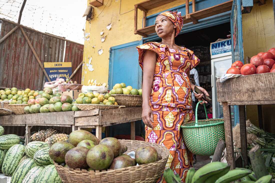 In this photo, Jeannette Uwimbabazi stands outside a store, where fruits are on display for sale outdoors.