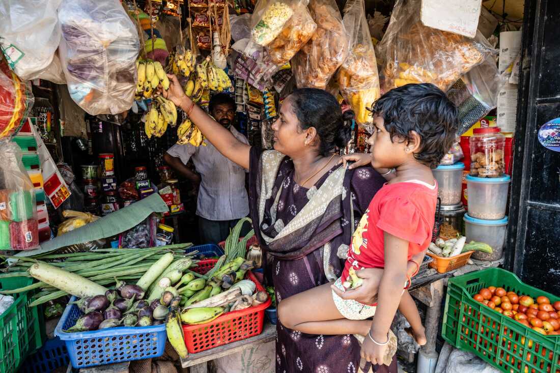 In this photo, Lakshmi is holding her daughter while standing outdoors in front of a grocery vendor. She's reaching for a banana that's hanging from a bunch. The vendor also has vegetables and other groceries on display for sale.