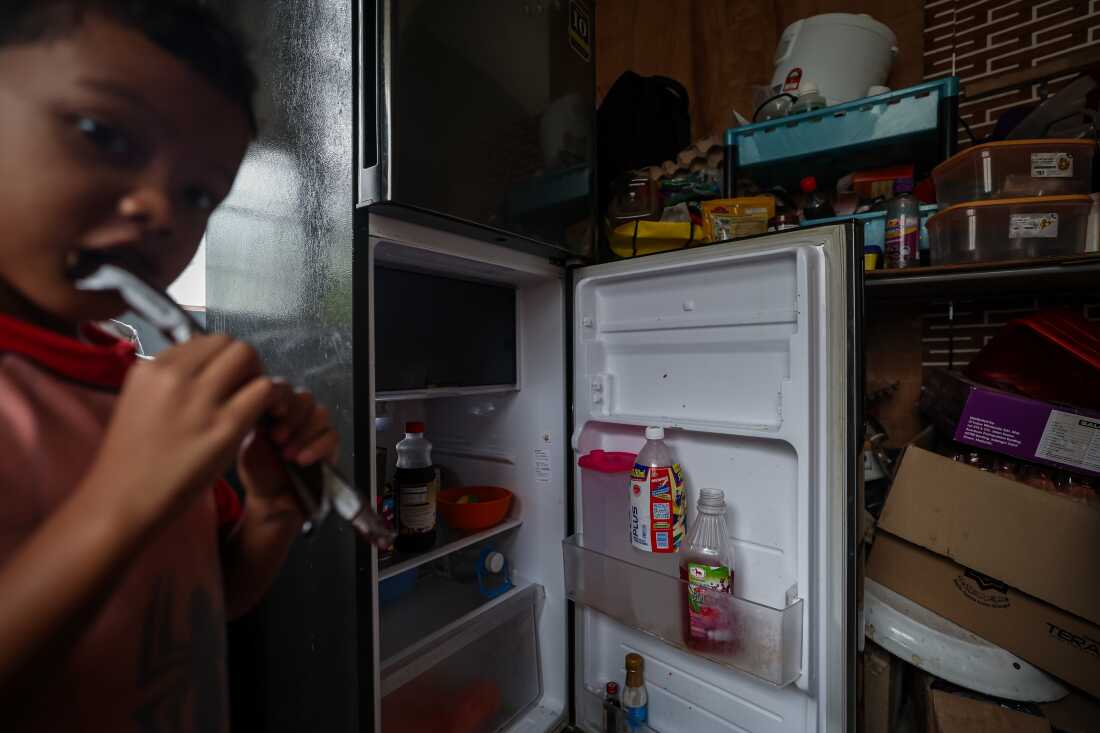 This photo shows the opened fridge in Rosnah's home. The fridge's contents are sparse; a bottle of juice and a bottle of water are in a shelf in the door. On the left, 5-year-old grandson Qayyum eats a chocolate waffle biscuit treat.