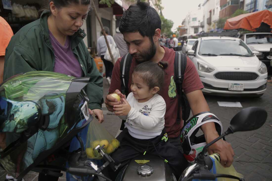 In this outdoor photo, Tomás sits in front of his father on his father's motorbike while reaching for a fruit that his father is holding. His mother stands near them, holding a bag of fruit. 