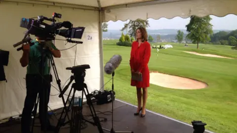 Andrea Byrne / Y Lolfa Andrea Byrne wearing a red dress stands in front of a camera and microphone ready to present the ITV Wales news