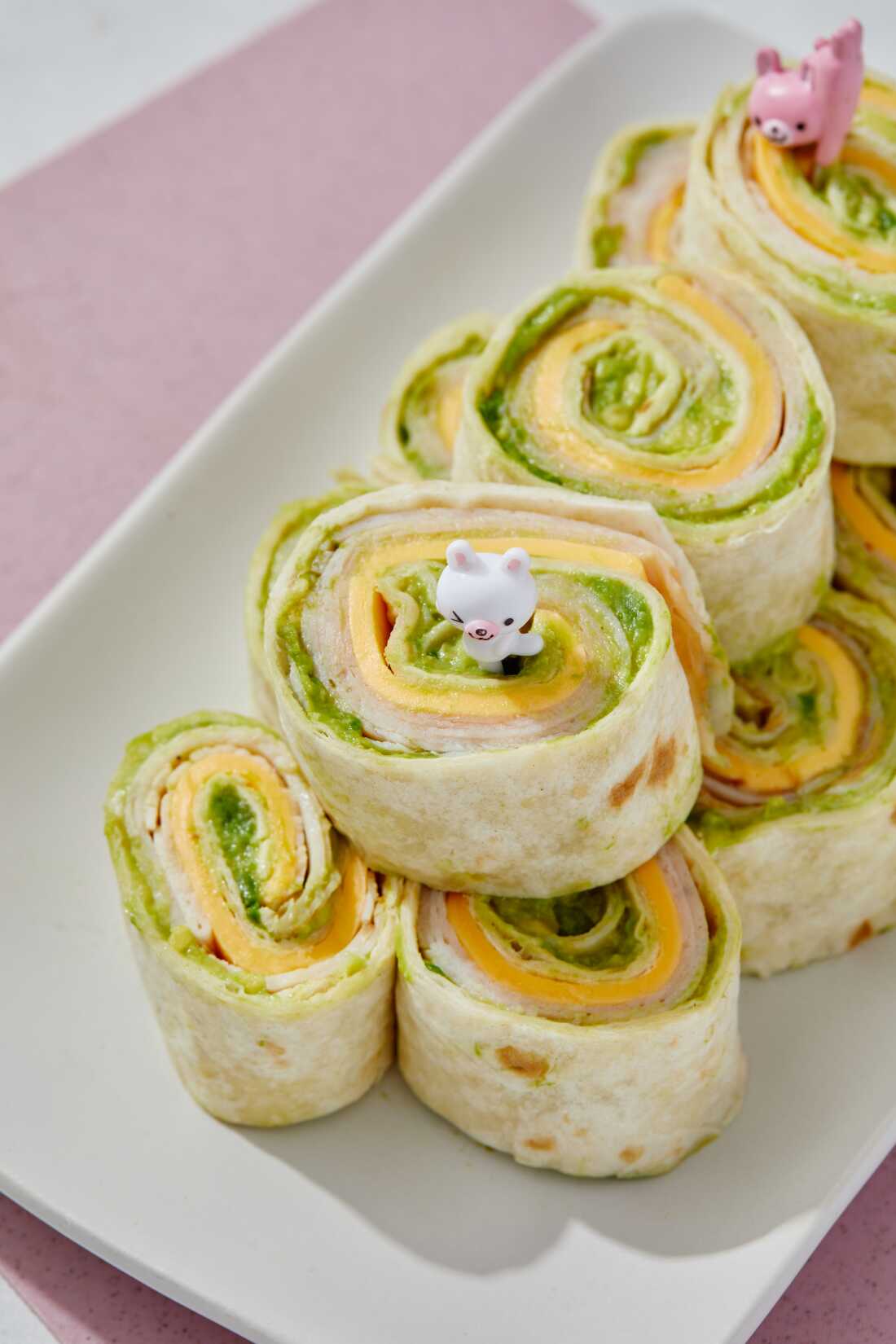 Photograph that shows turkey, avocado and cheese pinwheels stacked on a white plate.  Sliced turkey, cheese and avocado are layered in a large flour tortilla and rolled up tightly.