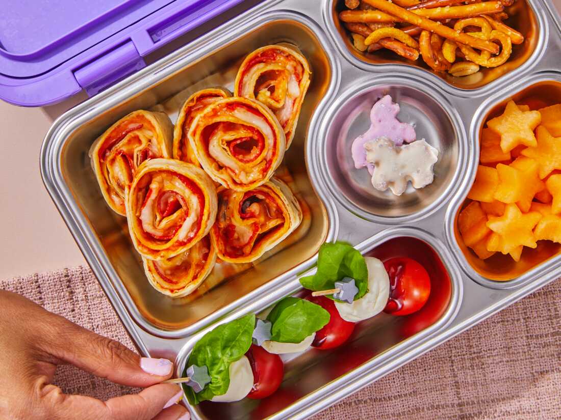 A high-angle photograph of a purple and metal bento box lunchbox that contains pepperoni and mushroom pizza roll-ups, caprese salad bites, pretzels and fruit. 