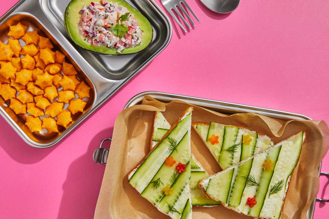 A high-angle studio photograph of an open-faced cucumber sandwich and shrimp-stuffed avocado in a metal lunchbox with compartments on a pink background. 