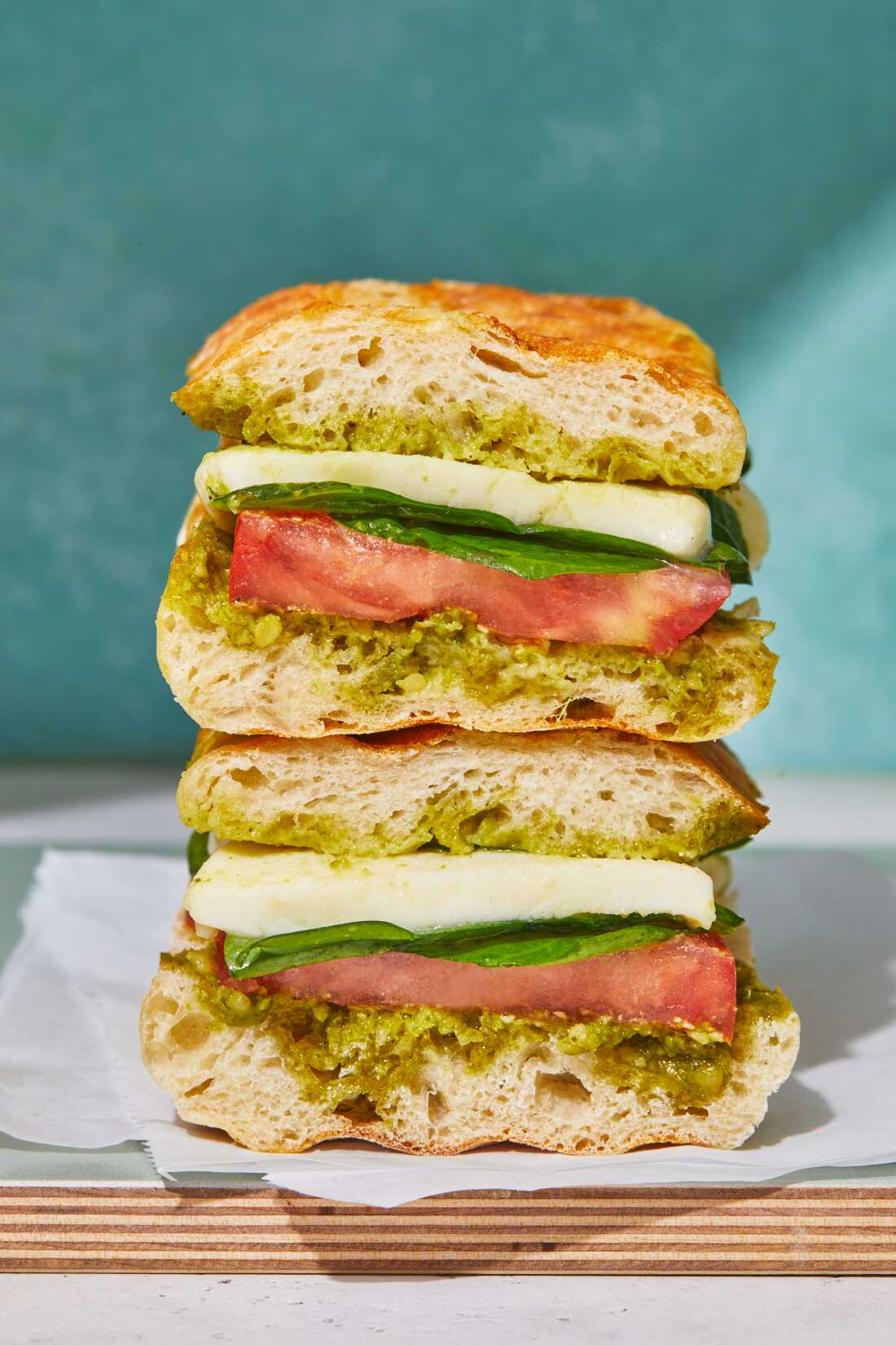 A close-up photograph of a mozzarella, tomato and spinach pesto panini cut in half and stacked on a wooden board in front of an aqua-colored background. 