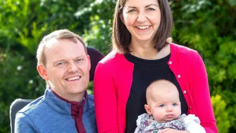 My Name'5 Doddie Foundation Scott Stewart sits in his wheelchair while his wife, Robyn, and daughter Rae are perched on the edge and smiling