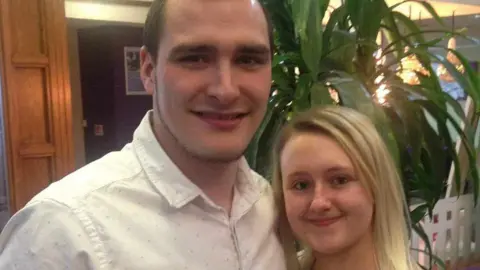 HANDOUT Smiling Chris Taylor, who has brown hair and a beard and is wearing a white shirt, is pictured with his partner Gemma, who has long blonde hair and minimal make-up, in the foyer of a venue