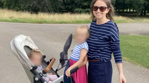 Family handout A woman in a park with two children. She has dark hair and is wearing sunglasses, a blue and white striped top, and blue trousers. The woman is standing in front of a buggy - there is a child standing on the back of the buggy who has blonde hair and is wearing a blue and white striped top and a pink skirt. Her face is blurred out. Sitting in the buggy is a toddler whose face is also blurred. Behind them is trees and grass.