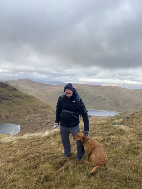 Glenn with his dog Ruadh on a grassy mountainside. Ridges and two small lochs can be seen in the distance and the sky is cloudy.