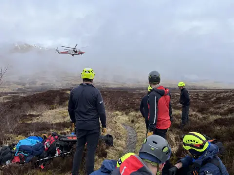 Five members of the rescue team with their backs to the camera on a hillside, watching a helicopter flying in the distance