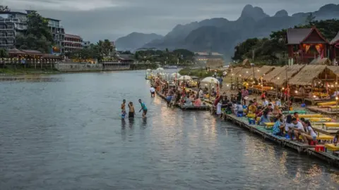 Getty Images Wide view of a river in Vang Vieng. Three people can be seen in the river while crowds are seen sitting along the river front. Mountains can be seen in the background with the town's skyline