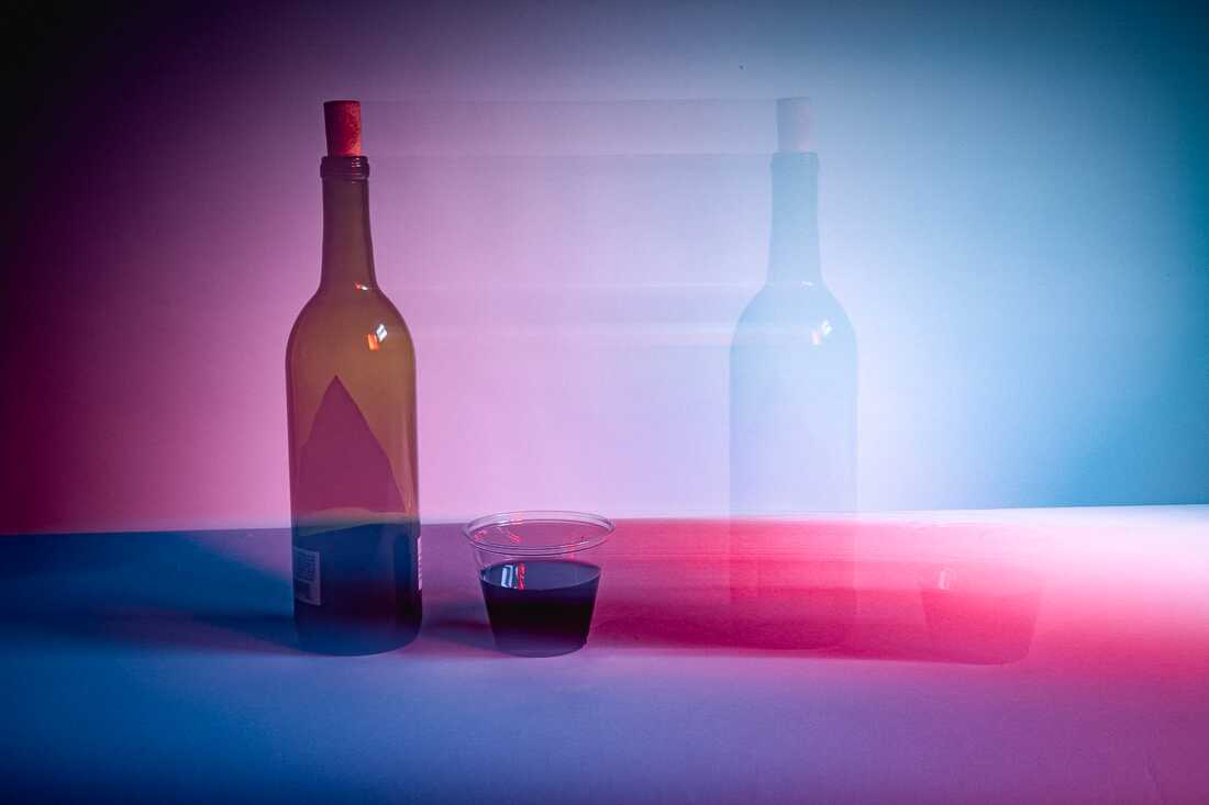 Photograph shows a bottle of wine and a plastic party cup filled with wine on a blue and purple backdrop with motion blur that puts the cup of wine and the bottle of wine to the right of the objects. 