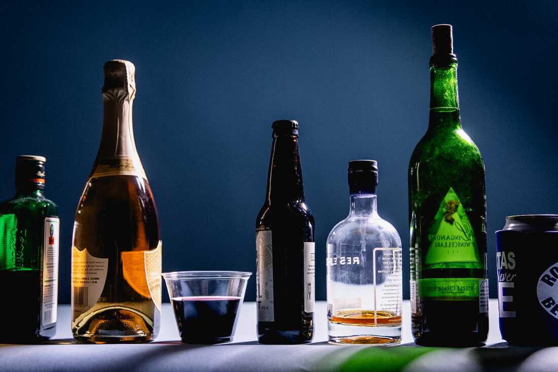 Studio photograph showing various kinds of alcohol bottles, cans and cups lined up in front of a blue background, including liquor, wine and beer. 