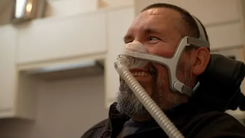 BBC A smiling Nik Ward looks towards his family who are standing to the left, out of shot. He has a beard and is sitting in a wheelchair with a breathing mask across his face.