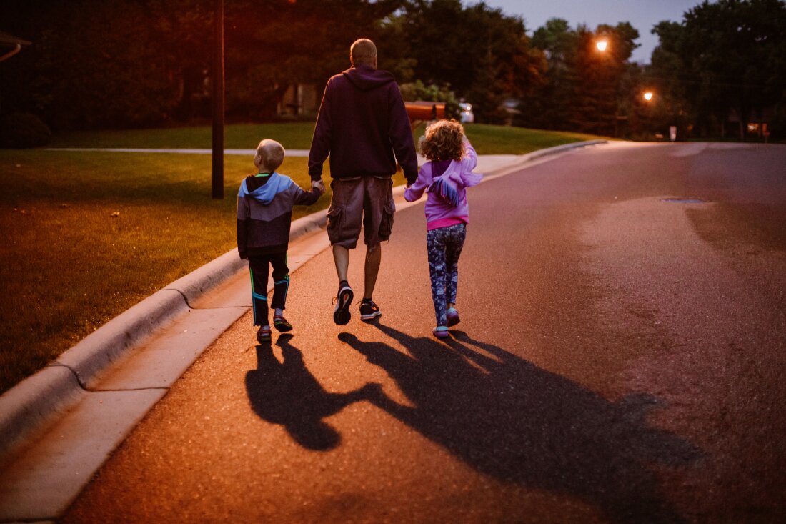 Photographed from behind, a man, a girl and a boy walk down a neighborhood street. The man is standing between the two children, holding the children's hands. The sky is getting dark as night appears to approach.