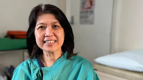 BBC Jennifer Loke sits in a doctor's consulting room with an examination couch behind her. She is smiling, has long black hair and is wearing green scrubs.