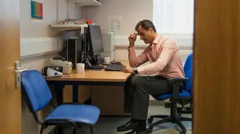 Getty Images A GP sits at his desk, holding his hand to his head, with an empty patient's chair beside him.