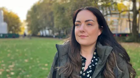 BBC Photo of 31-year-old lady. She is looking into the distance and has a winter coat on. She is outside on grass, with a row of trees in the distance behind her. 