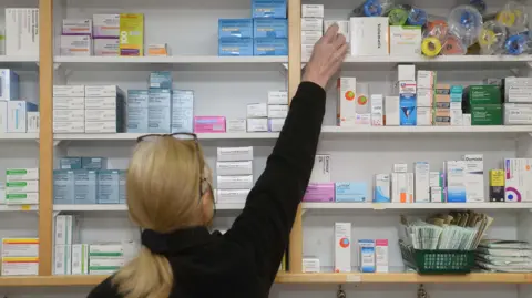 MARK MARLOW/EPA-EFE/REX/Shutterstock The photo shows a woman with blonde hair tied in a low ponytail, with glasses on her head, wearing a black jumper. She is facing away from the camera and reaching onto the top shelf of a wall full of different medications.

