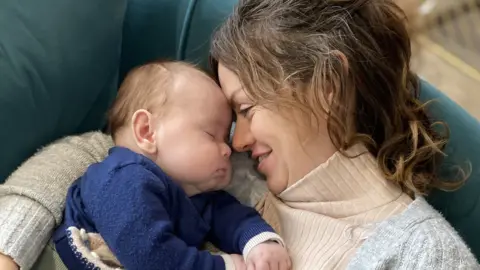 Family handout Image of a woman cuddling a sleeping baby. She has her hair tied back, is wearing a roll neck jumper and is smiling. She has her faced pressed against the baby's, which has its eyes closed. The baby is wearing a dark coloured jumper and has very chubby cheeks.