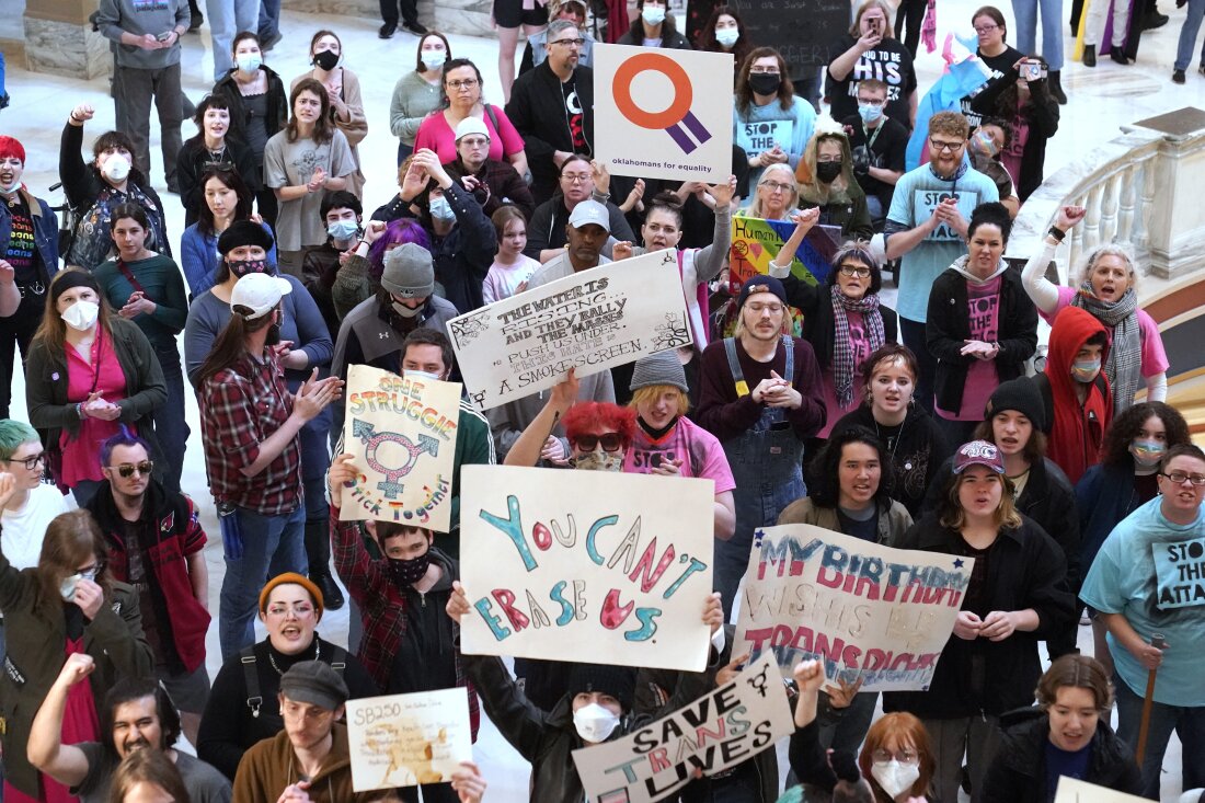 A crowd of people are photographed from above in the Oklahoma state capitol. Some of them hold handmade signs supporting trans rights. A sign in the middle reads, "You can't erase us."