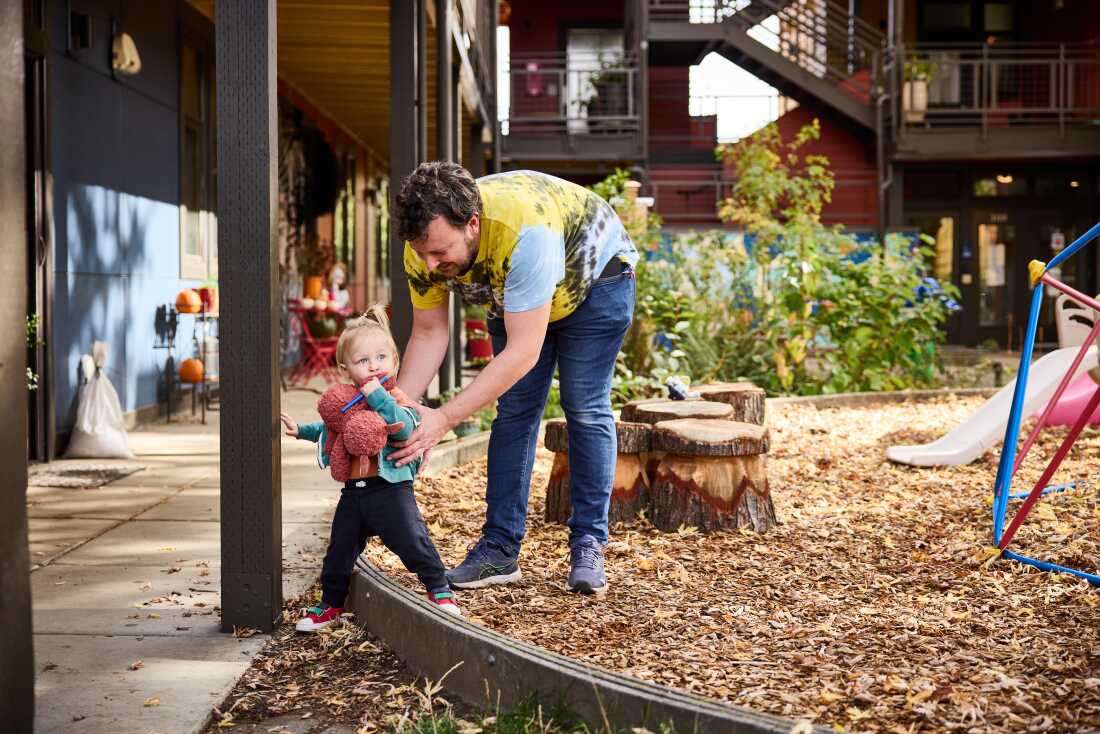 A dad helps his toddler navigate a low playground barrier.  The dad is wearing a tie-dye t-shirt; the little girl holds a red stuffed-animal toy.