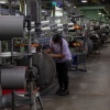 Alexandra Vazquez works in the Phifer factory, a window screen manufacturer outside Tuscaloosa, Alabama. A woman in a purple t-shirt inspects a large spool on a factory floor with many more large spools of material. 