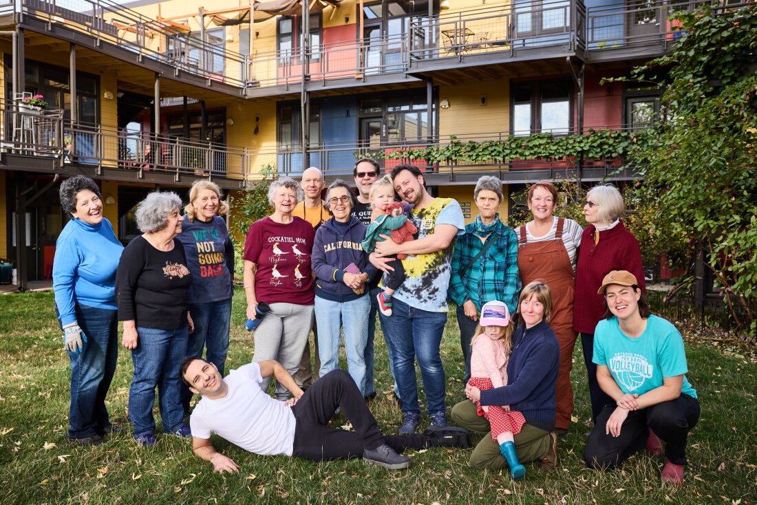 Sixteen people, including two small children and several older people, stand in a grassy courtyard in front of a colorful modern building. Most of them have big smiles on their faces. 