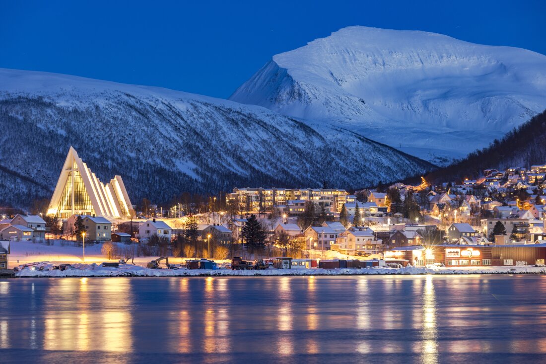 Dusk falls over snowy mountains overlooking the Arctic Cathedral and houses mirrored in the icy fjord in Tromsø, Norway. Although this city is north of the Arctic Circle and experiences polar night for two months out of the year, its residents tend to see winter as a time of opportunity.