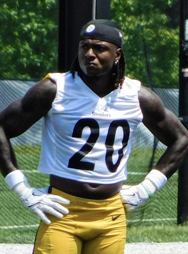 A football player wearing a white practice jersey with the number 20, yellow pants, gloves, and a black head covering stands outdoors with his hands on his