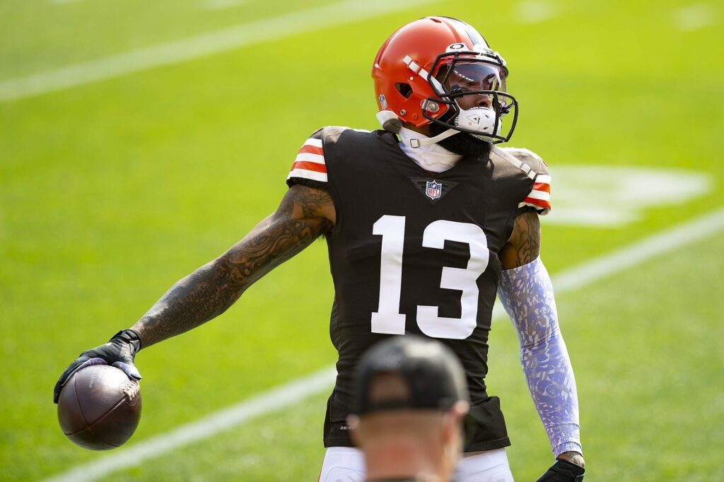 A football player wearing a black number 13 jersey and orange helmet holds a football on a field, with another person's head partly visible in the foreground.