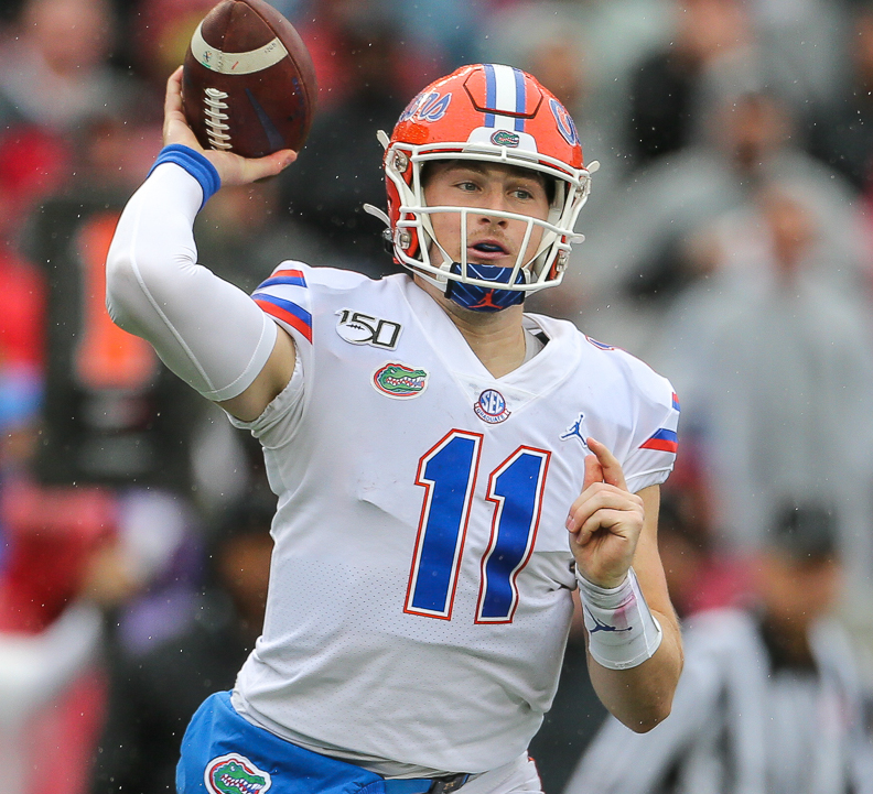 A helmeted American football player in a white uniform with blue and orange accents, number 11, holding a football in a throwing motion on a field.