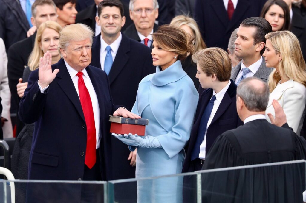 Donald Trump standing among officials and family members at a swearing-in ceremony, with a woman holding a book beside him.