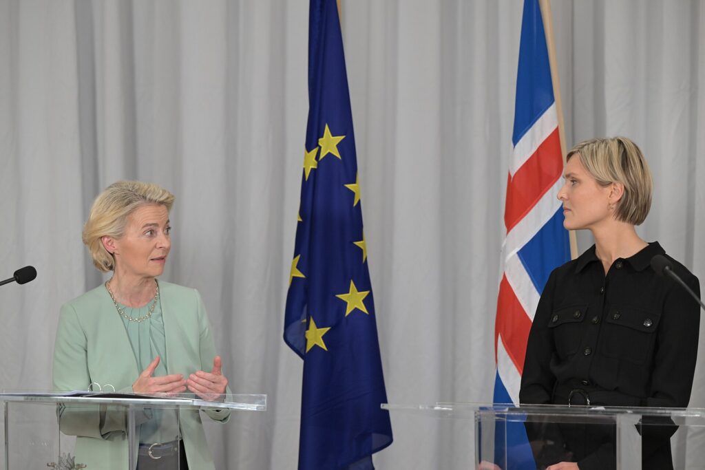 Two women stand at separate clear lecterns in front of European Union and Icelandic flags, with microphones and white curtains behind them.